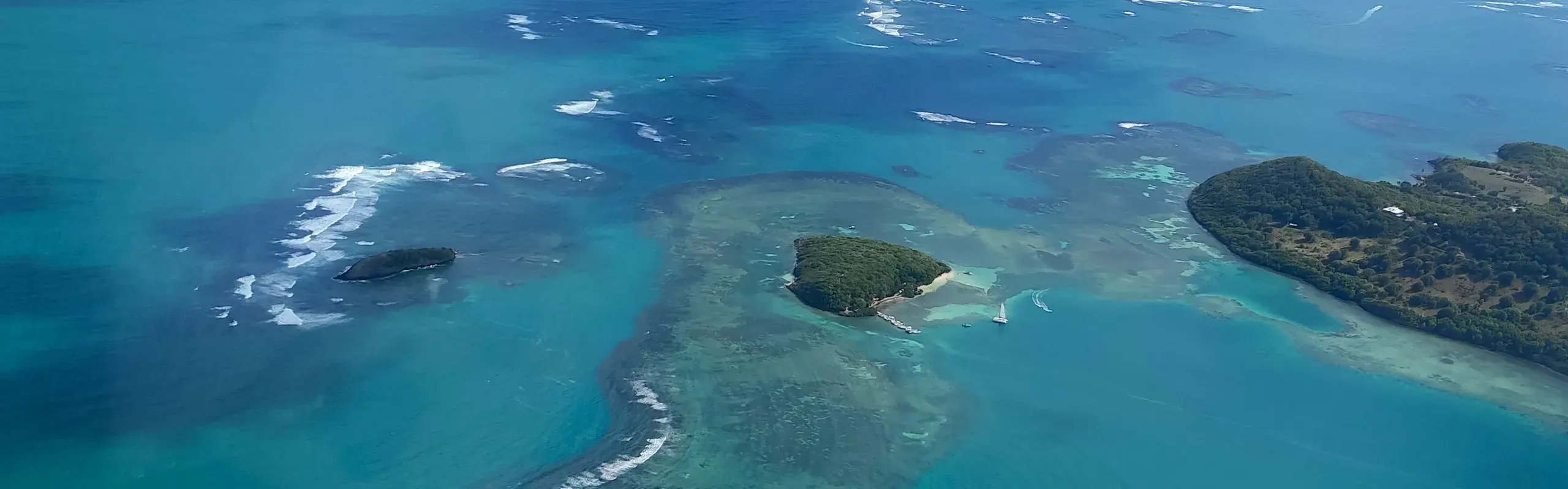 Vue aérienne de la baie du Robert : îlets boisés, catamarans au mouillage et récif où déferle la houle